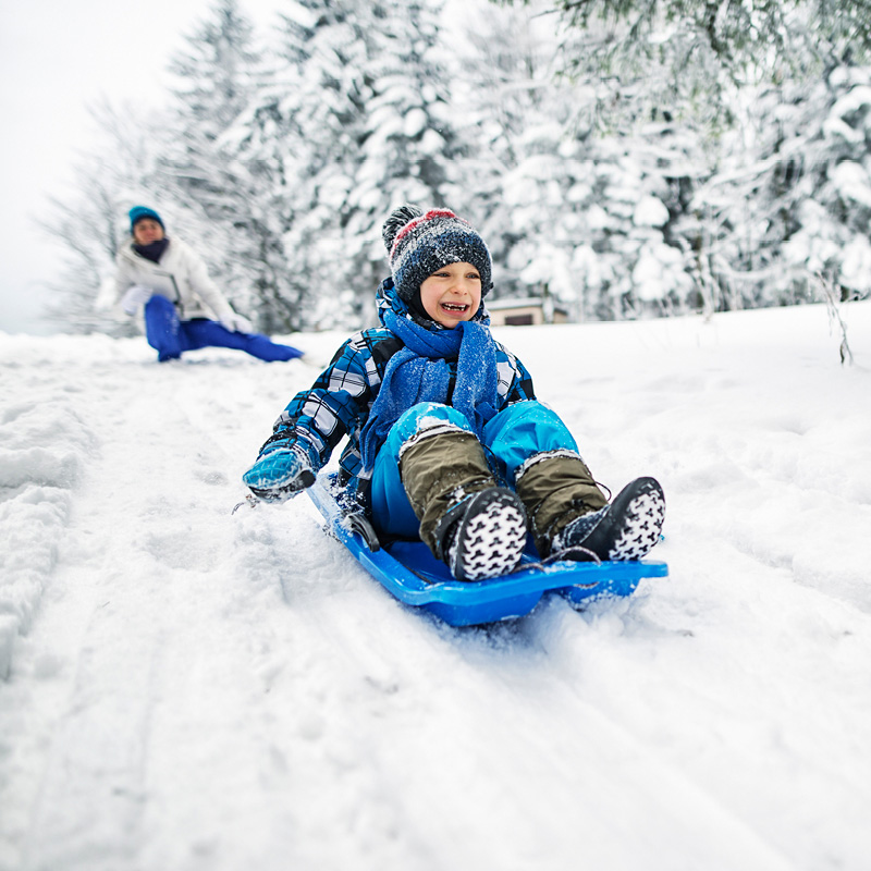 Lapsi sinisellä pulkalla vyöryy lumisessa talvimaisemassa, taustalla lumiset kuuset