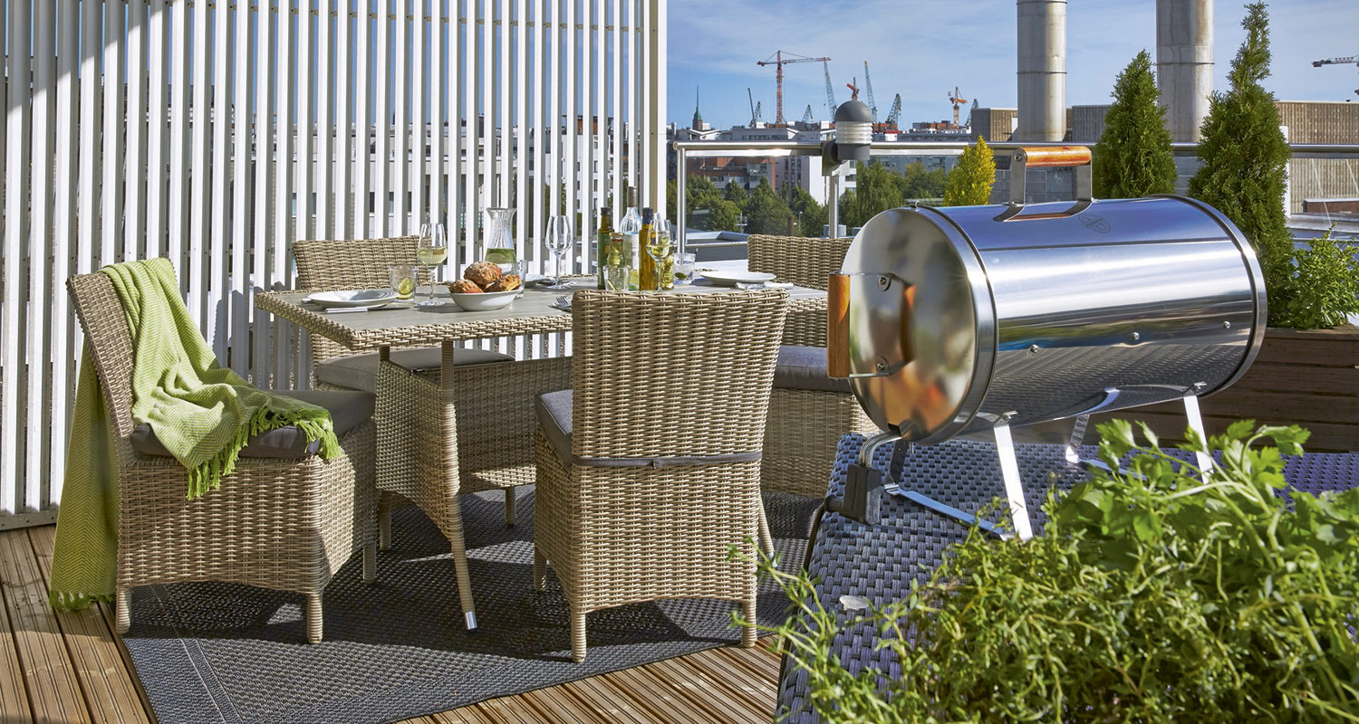 Aurinkoinen kattoterassi, rottinkikalusteet katetulla pöydällä, kiiltävä hiiligrilli ja kaupunkitausta.