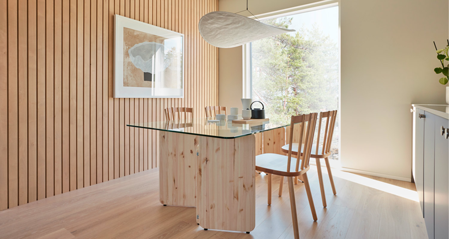 Valoisa ruokailuhuone: puupaneeliseinä, lasipöytä puupilarilla ja neljä vaaleaa puutuolia.