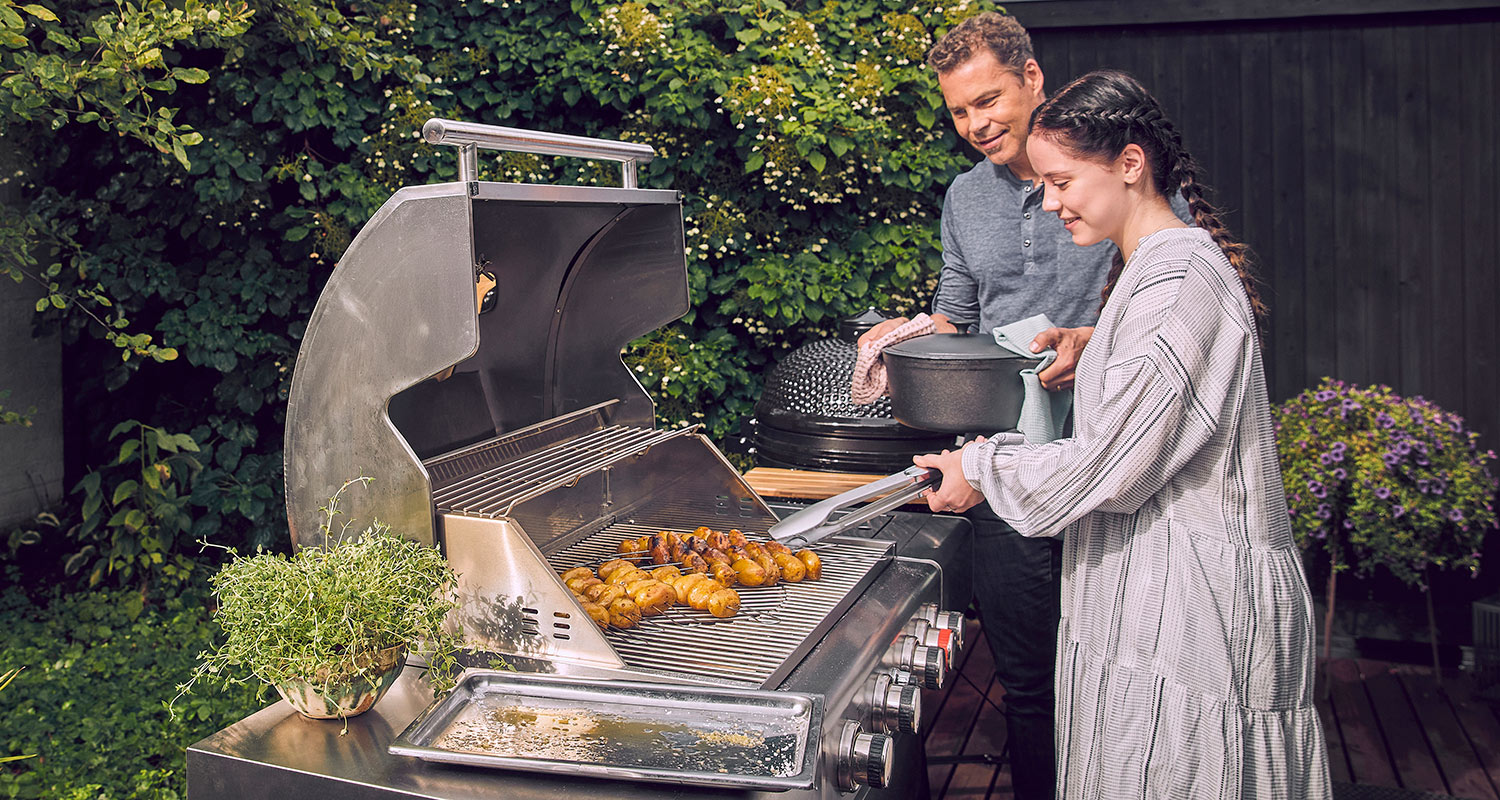 Nainen grillaa teräksisellä kaasugrillillä pieniä kultaisia perunoita vehreässä puutarhassa.