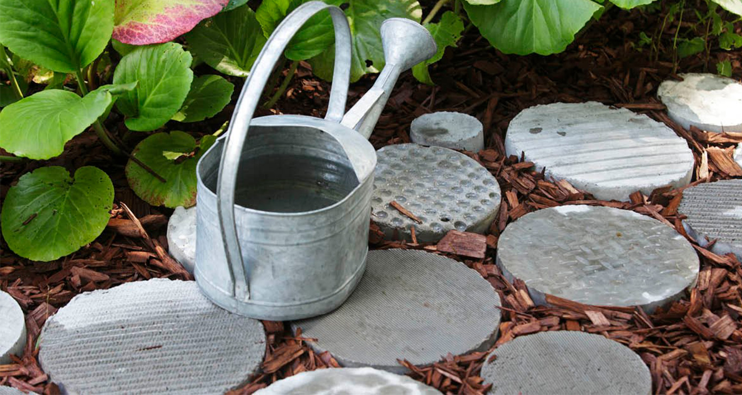 Metallinen kastelukannu pyöreillä betonilaatoilla ruskeassa hakkeessa ja vihreissä lehdissä