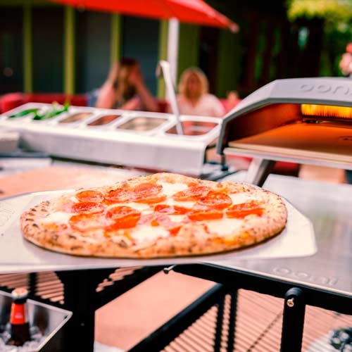 Pepperonapizza ulkoilmapizza-uunissa, taustalla täytesäiliöitä ja punainen aurinkovarjo