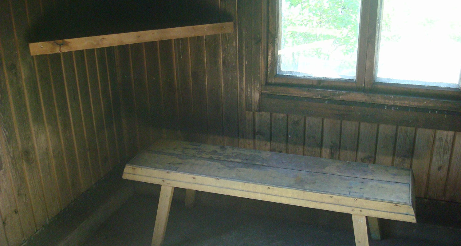 Sisätila hirsiseinillä, yksinkertainen vaalea puupenkki ja ikkuna, valoa tulvii sisään.