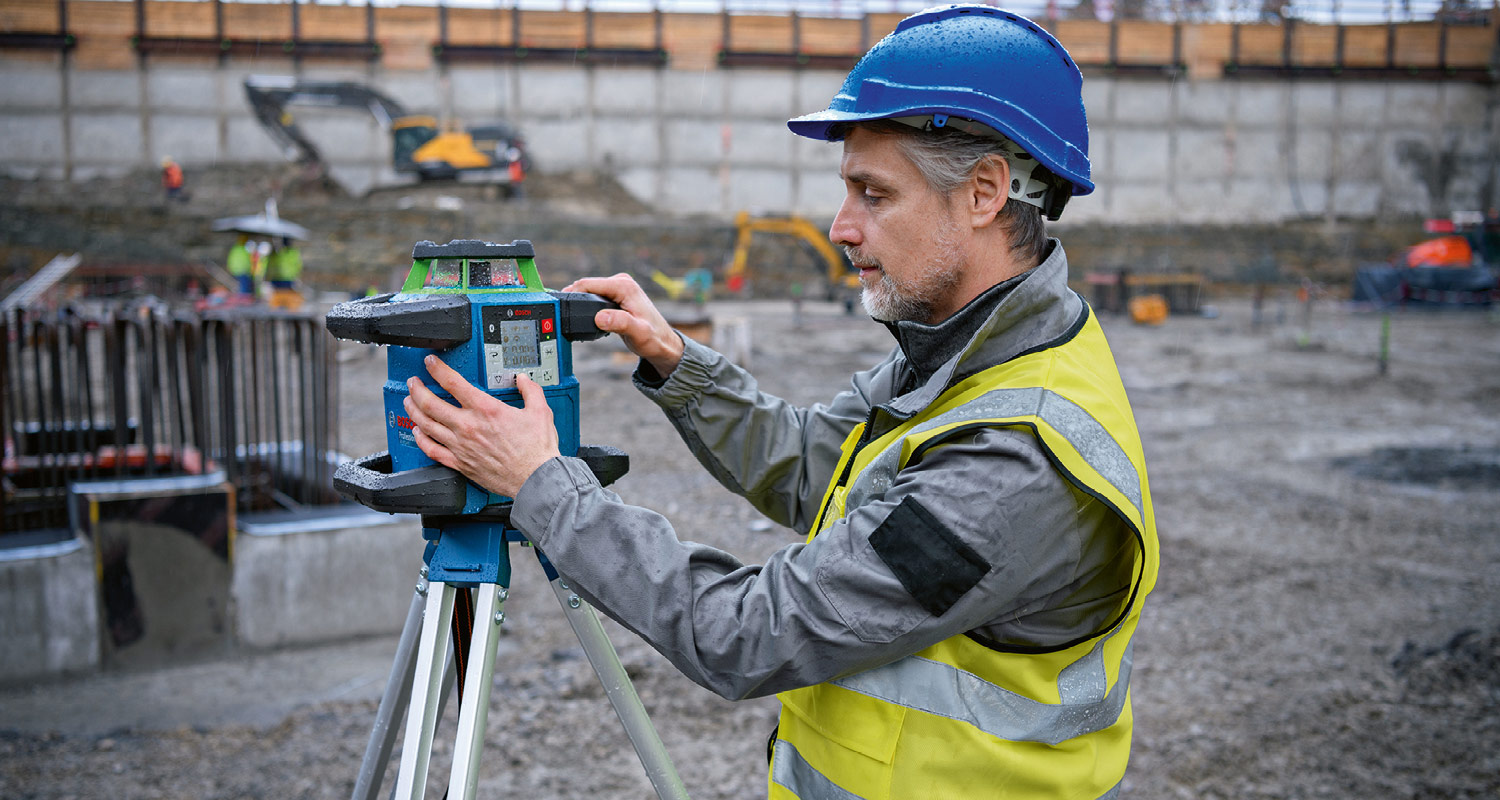 Rakennustyömaalla turvaliivinen työntekijä säätää jalustalla olevaa sinistä kolmiomittauslaitetta