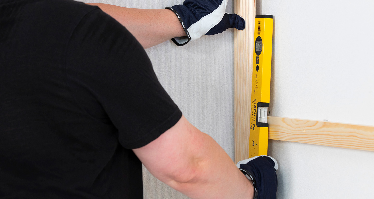 Mustapaitainen suojakäsineissä mittaa keltaisella vesivaakalla puurimaa valkoisella seinällä