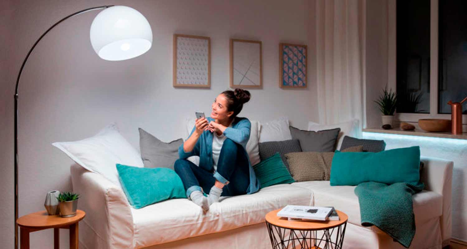 Henkilö selaa älypuhelinta valkoisella kulmasohvalla turkoosin ja harmaan sävyisillä tyynyillä