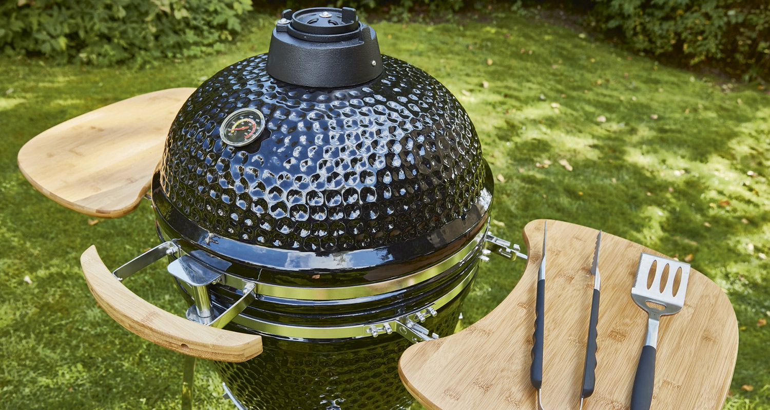 Mustakeraaminen kamado-grilli puisilla sivuhyllyillä, grillivälineet ja vihreä nurmikko.
