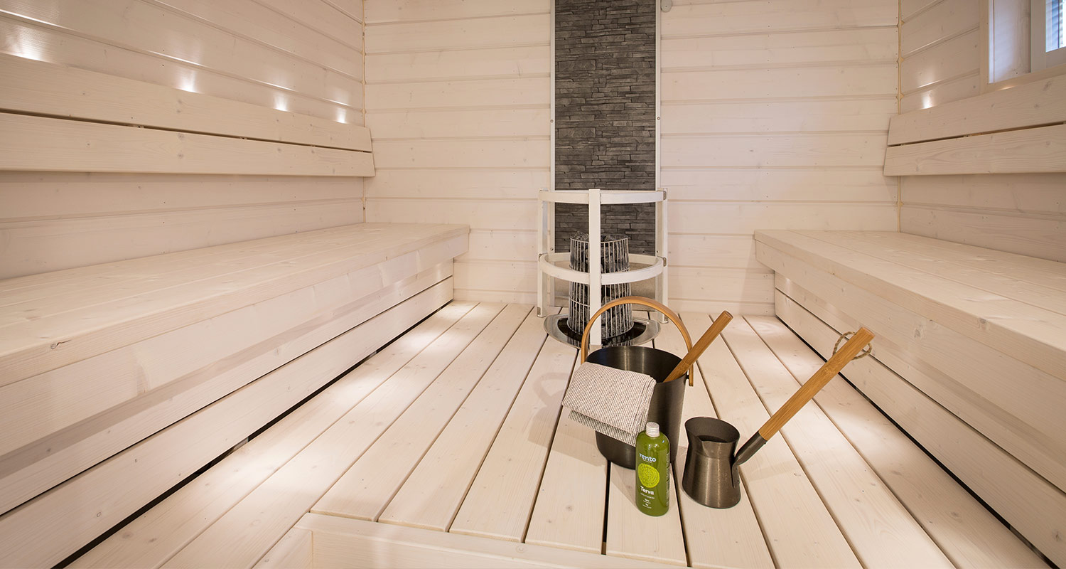 Vaalea moderni puusauna, keskellä kiuas, lattiolla musta löylykiulu ja vihreä tervapullo.