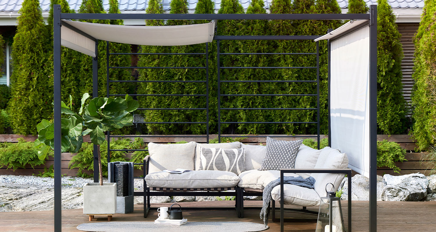 Terassi sisustettu K-Raudan Cello-terassikaluisteilla. Kuvassa sohva, pöytä, nojatuoli sekä pehmusteita. Kalusteet ovat metallisen ja tumman pergolan sisällä.