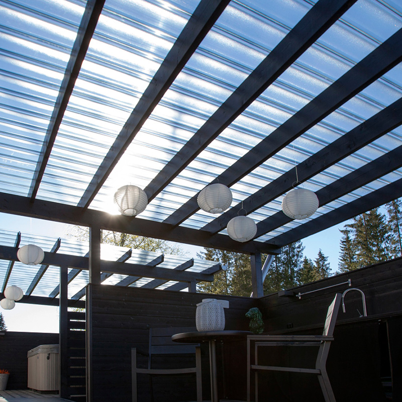 Aurinkoinen terassi metsämaisemassa, mustat puupalkit, läpinäkyvä katto ja valkoiset paperilyhdyt.