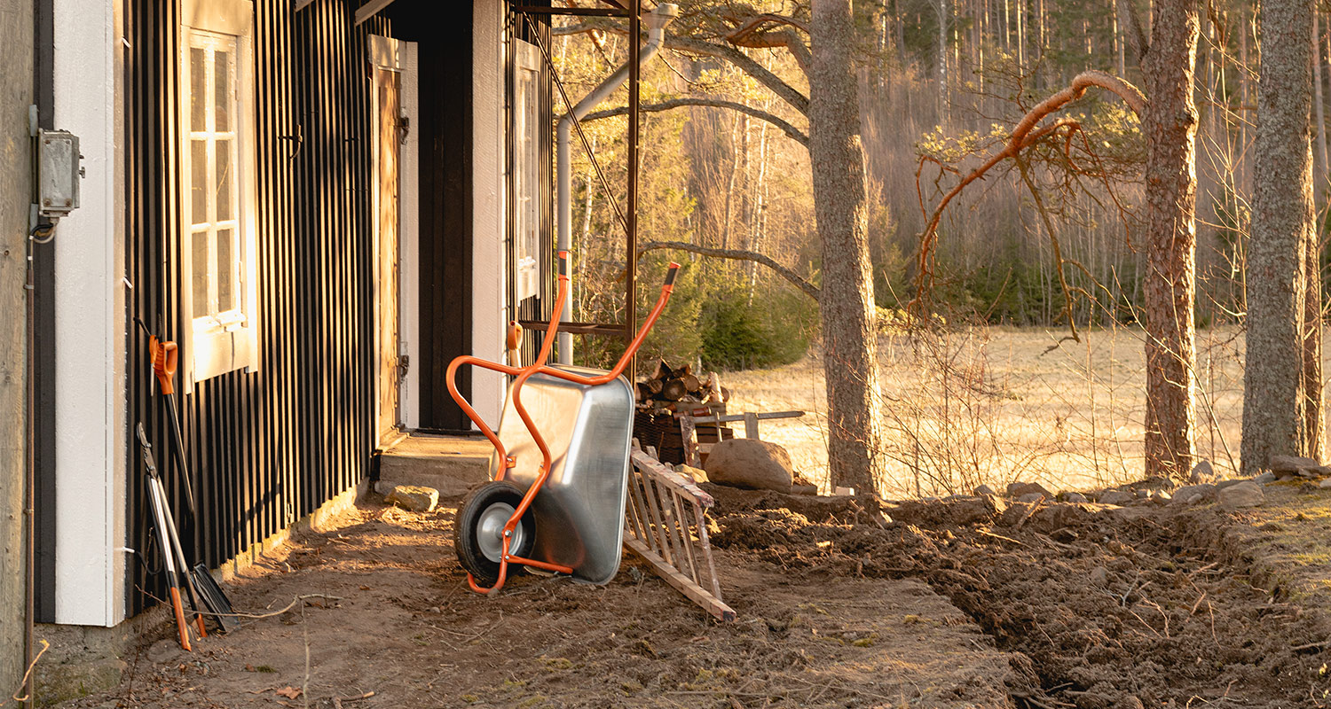 Oranssikahvainen kottikärry mudaisella pihalla mustan mökin ja havumetsän reunassa
