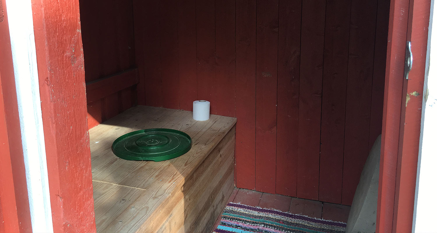 Punaisen ulkokäymälän sisällä puupenkki vihreällä kannella, vessapaperirulla ja räsymatto.