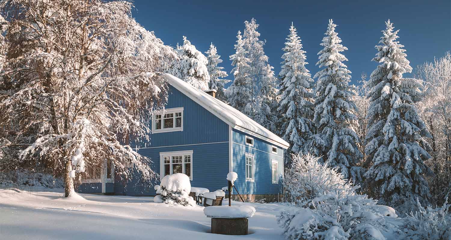 Sininen puutalo lumipeitteisessä metsämaisemassa, lumiset kuuset ja koivu kirkkaalla talvipäivällä.