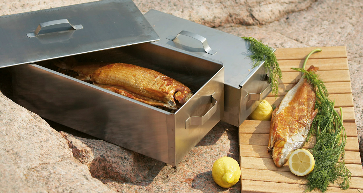 Avattu metallinen savustuskotelo kullanruskealla kalalla, vieressä puulaudalla sitruuna ja tilli.