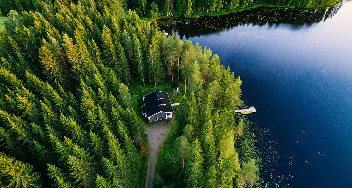 Ilmakuva tummakattoisesta mökistä tiheässä vihreässä kuusikossa järven rannalla laiturin vieressä