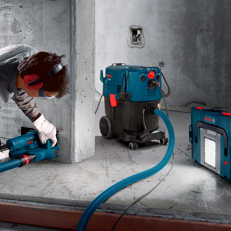 Rakennustyömies poraa betoniseinää sinisellä Bosch-porakoneella kiinni pölynimuriin ja valaisimeen