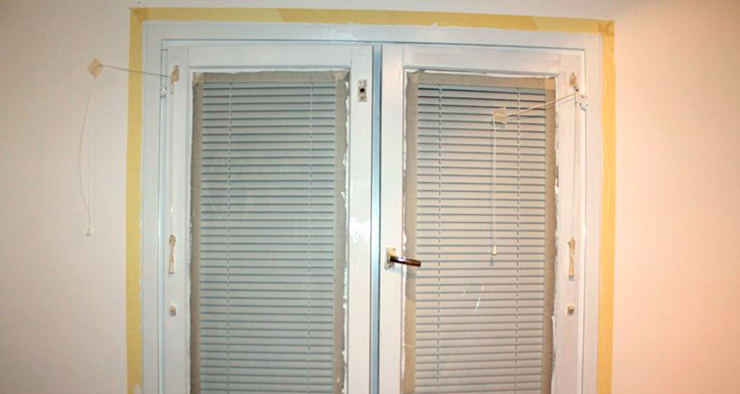 Valkoinen muovi-ikkuna keltaisella suojateipillä, sisällä suljetut harmaat sälekaihtimet.