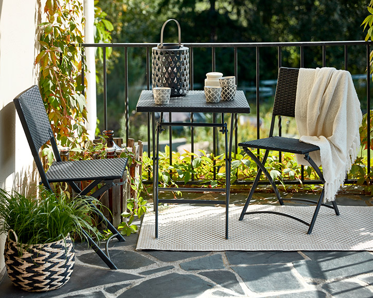 Parvekkeella mustat punotut tuolit ja pikkupöytä, kynttilälyhty, valkoinen viltti, vihreät köynnökset