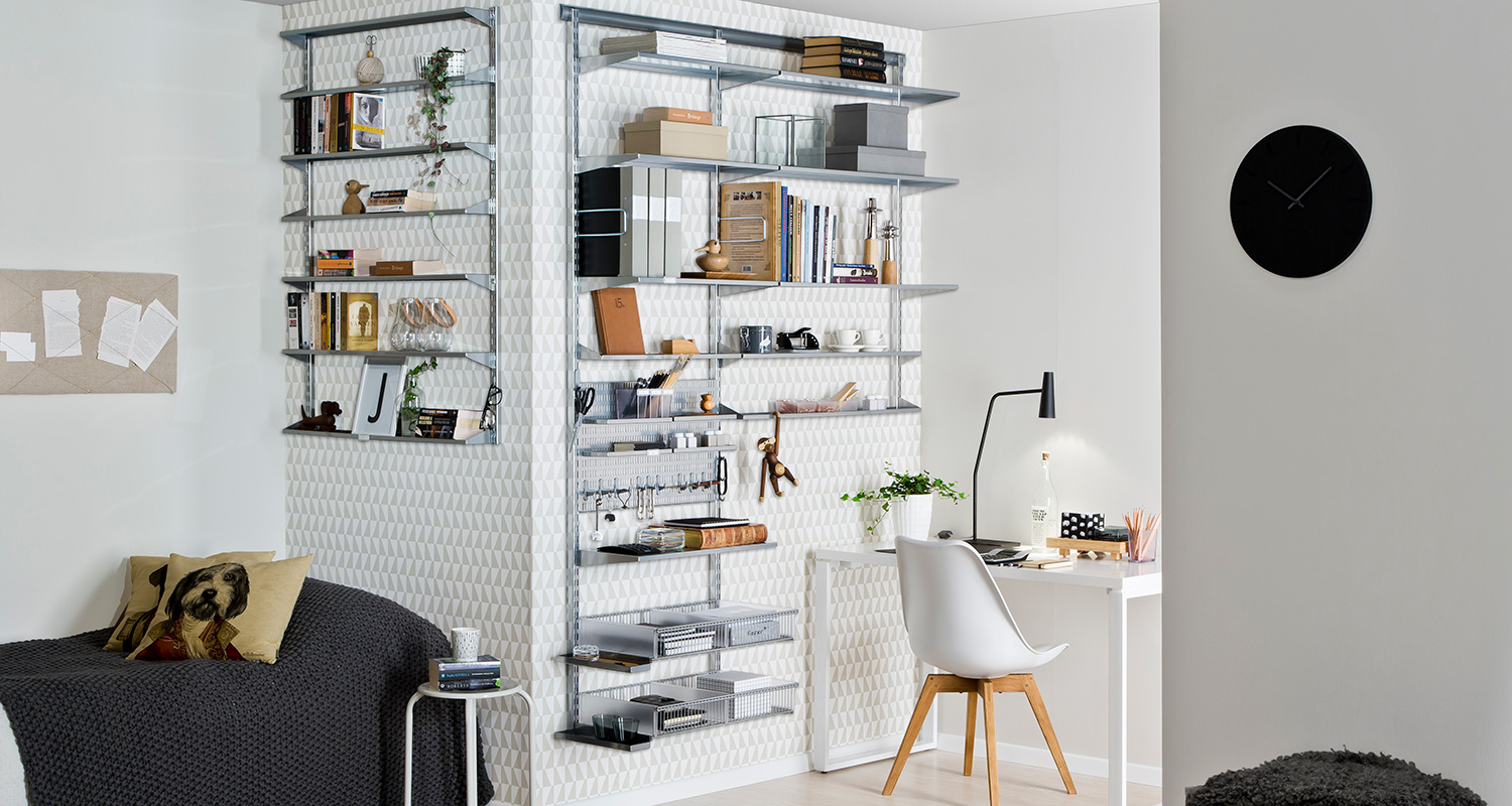 Skandinaavinen kodintyöpiste valkoisilla hyllyillä, viherkasvi, musta kello ja harmaa sohva.