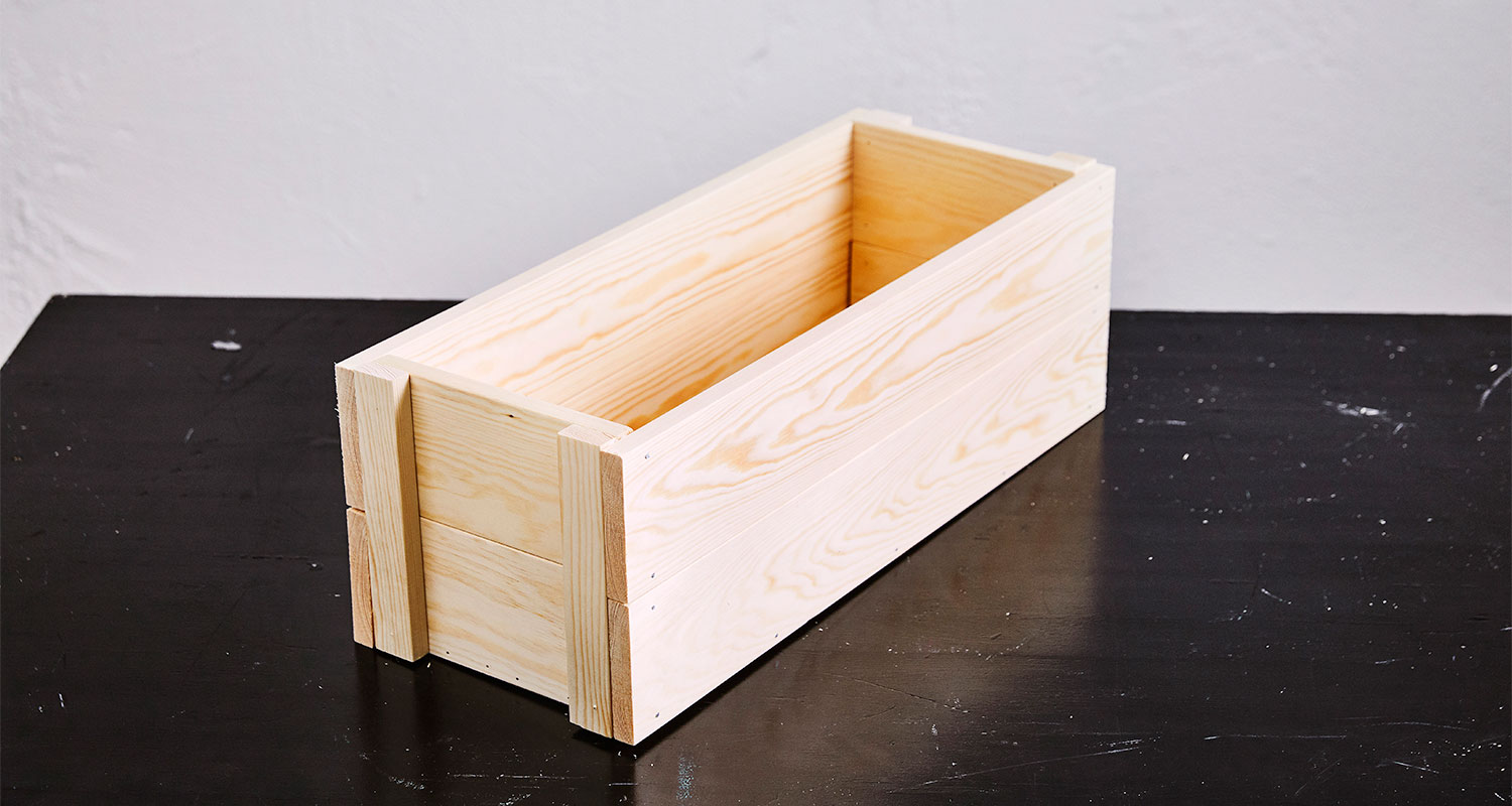 Pitkä vaalea puinen laatikko mustalla pöydällä valkoista seinää vasten