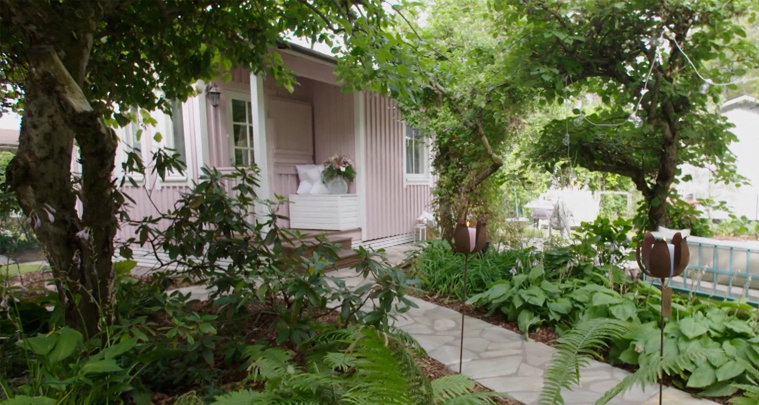 Vaaleanpunainen puutalo, vihreä köynnöskaari ja puutarha kivilaatta­polulla