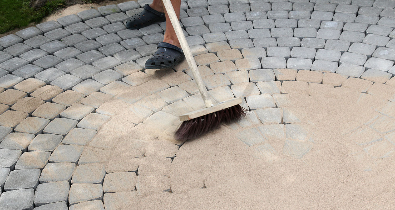 Kumitöppösillä seisova henkilö harjaa beigehiekkaa harmaiden, pyöreiden pihakivien väleihin