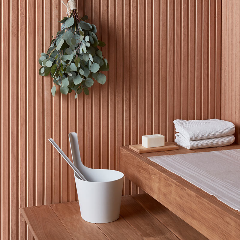 Saunan puupaneeliseinä, ripustettu eukalyptus, valkoinen löylykiulu, pyyhkeet ja saippua.