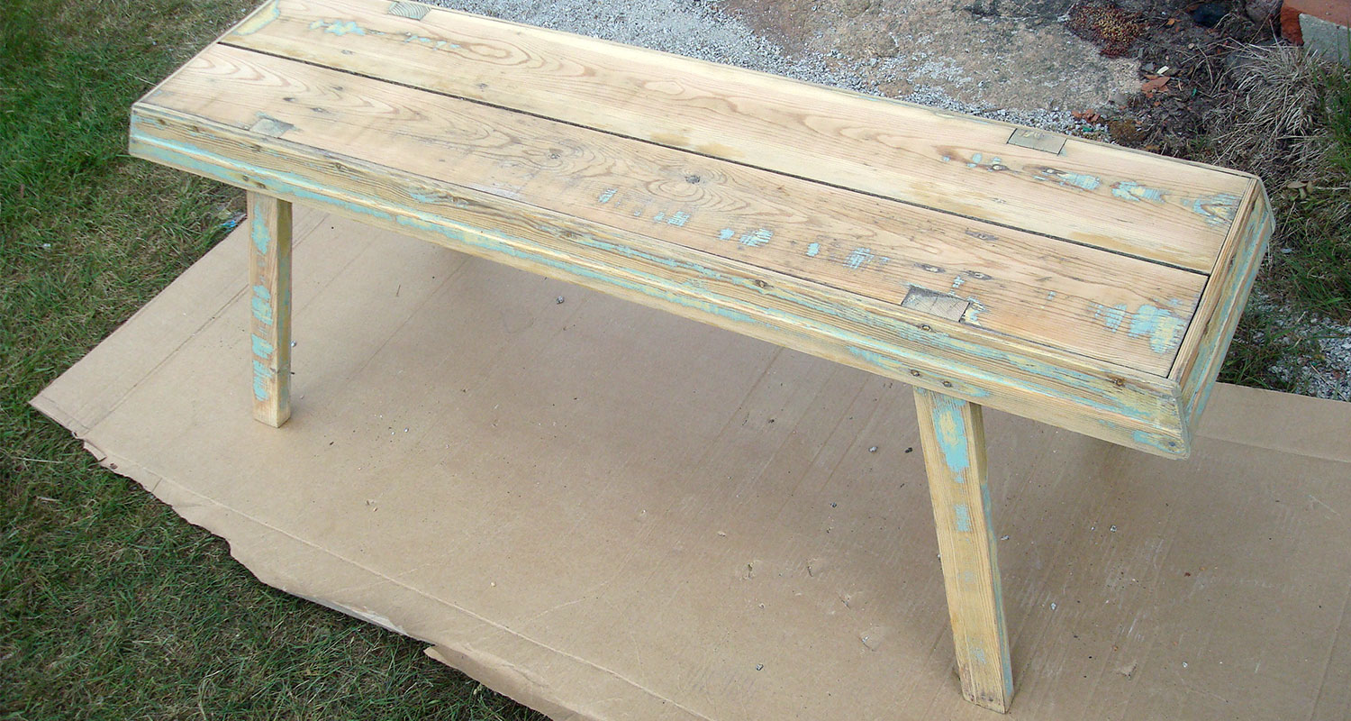 Haalistuneen turkoosilla maalilla pilkottu luonnonvärinen puupenkki pihalla kartongin päällä.