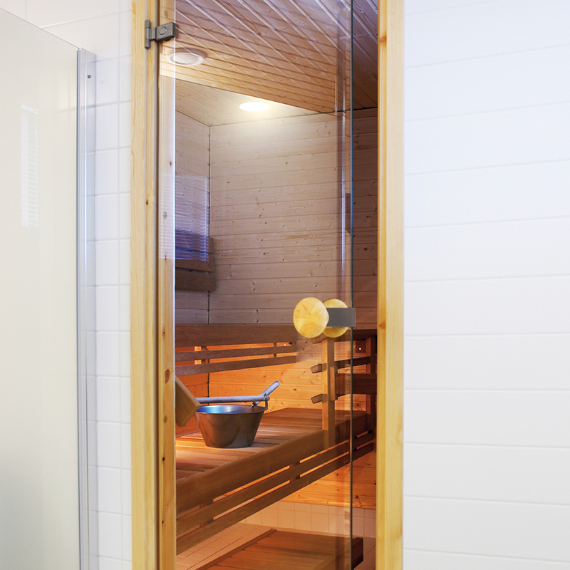 Lasiovinen saunahuone, vaalea puupaneeli, lämmin oranssi valaistus, kiulu ja kauha lauteilla