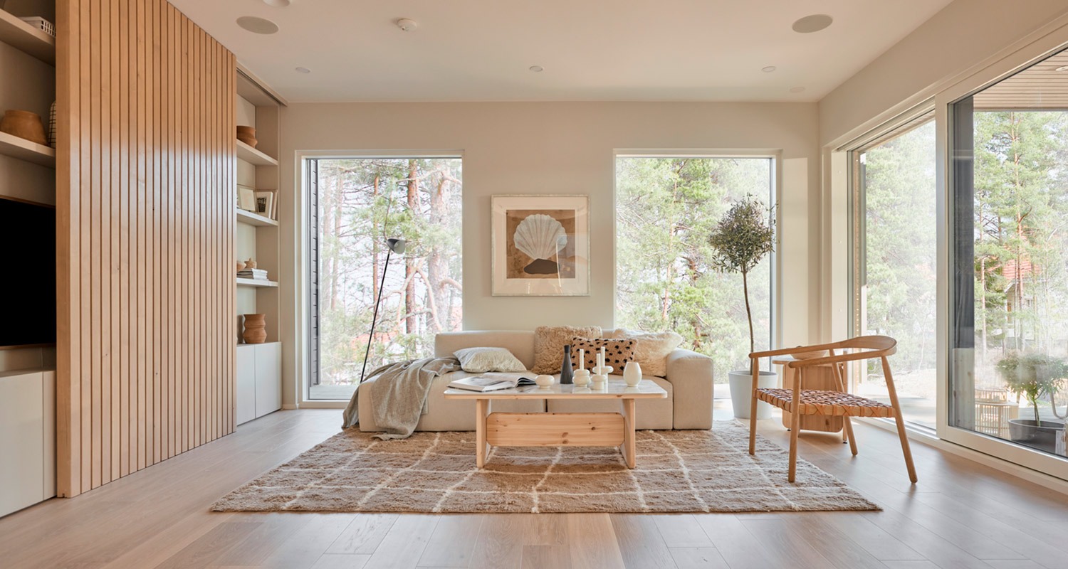 Moderni vaalea olohuone isoilla ikkunoilla, beige sohva, puupöytä ja nojatuoli metsänäkymällä.