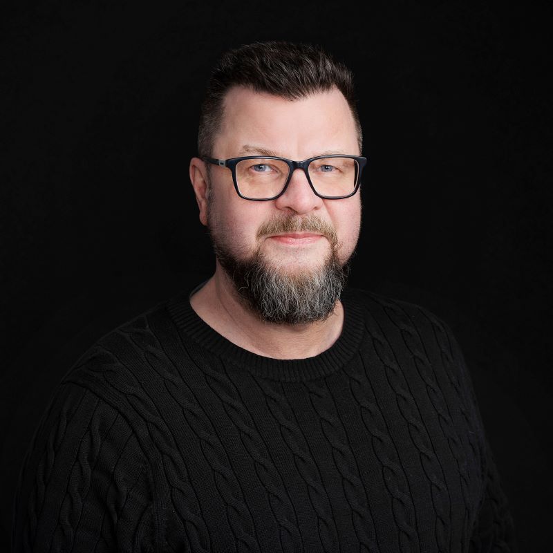 Miehinen ylävartalon muotokuva mustassa palmikkoneuleessa, harmaa parta ja tummanpuhuva tausta.