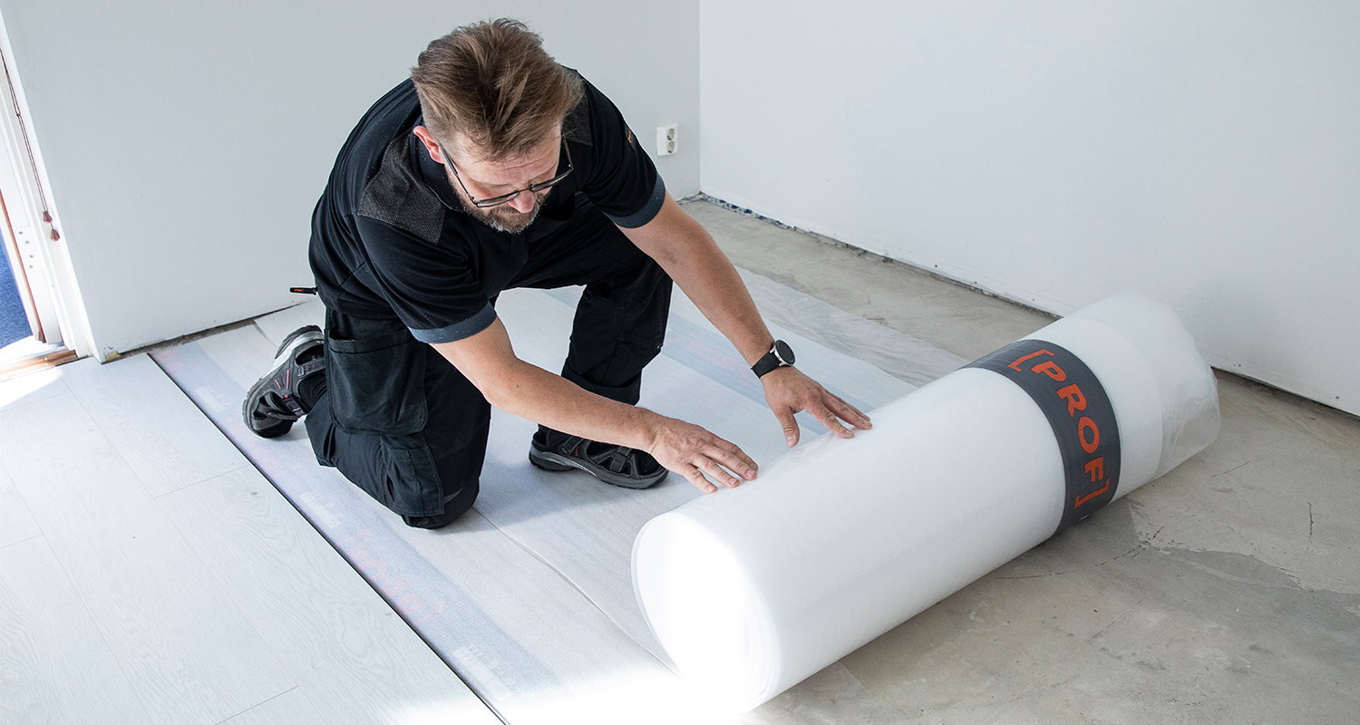 Työmies mustissa vaatteissa rullaa valkoista aluskumia vaalean laminaatin alle harmaassa huoneessa