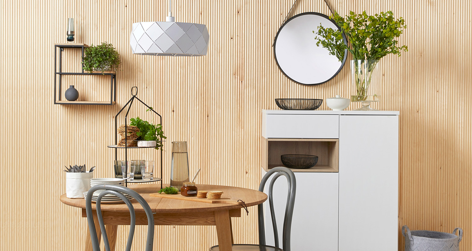 Vaalea puupaneeliseinä, pyöreä pöytä, harmaat tuolit, valkoinen lipasto, pyöreä peili ja viherkasvi