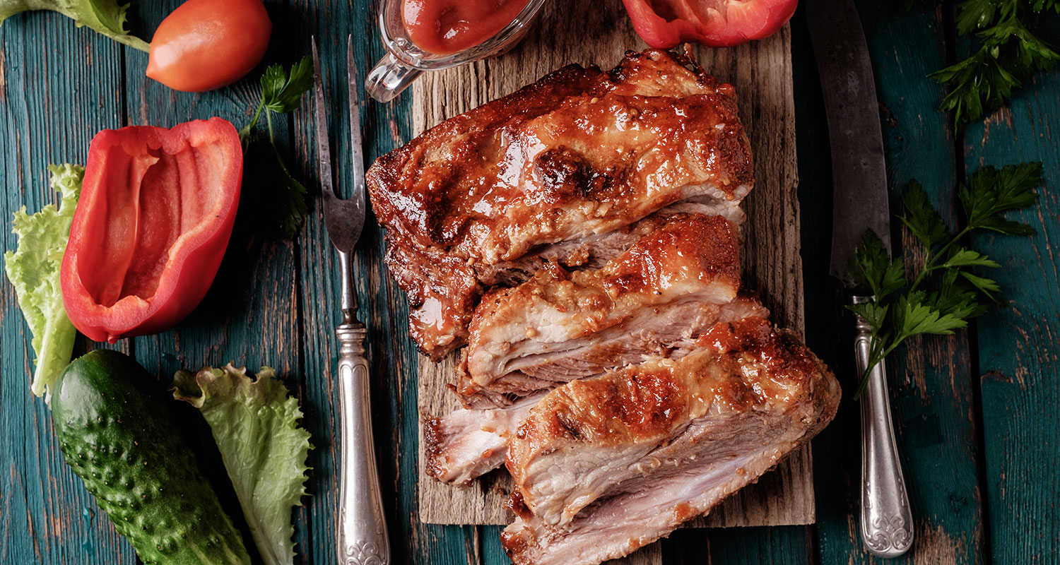 Glaseeratut grillatut porsaan kylkiluut puisella laudalla, paprikaa, kurkkua ja salaattia.