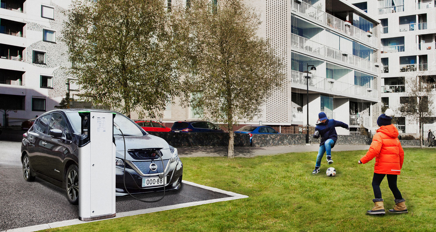 Musta sähköauto latauspisteessä kerrostalon edustalla, kaksi lasta pelaavat jalkapalloa nurmella.