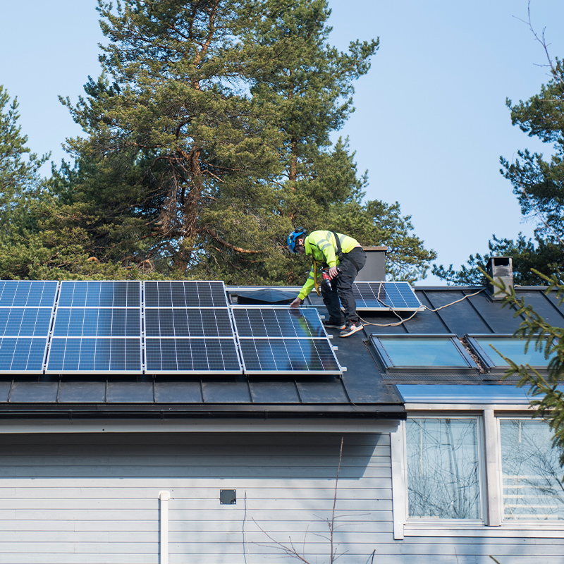 Kypäräpäinen työmies asentaa katolla aurinkopaneeleita mustan katon ja mäntyjen taustalla.