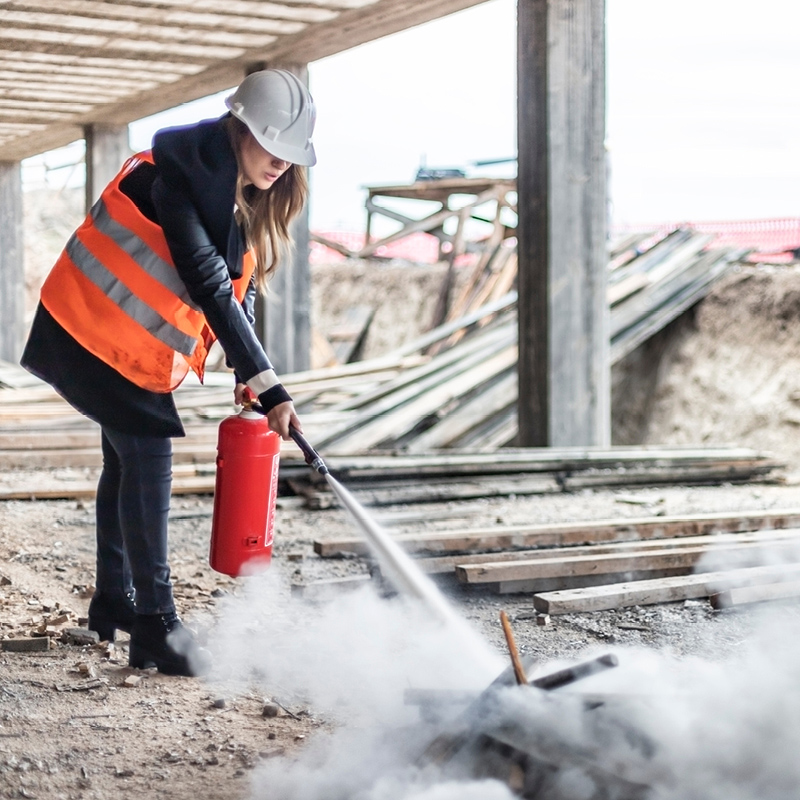 Rakennustyömaalla oranssiliivinen henkilö kypärä päässä sammuttaa savua punaisella sammuttimella