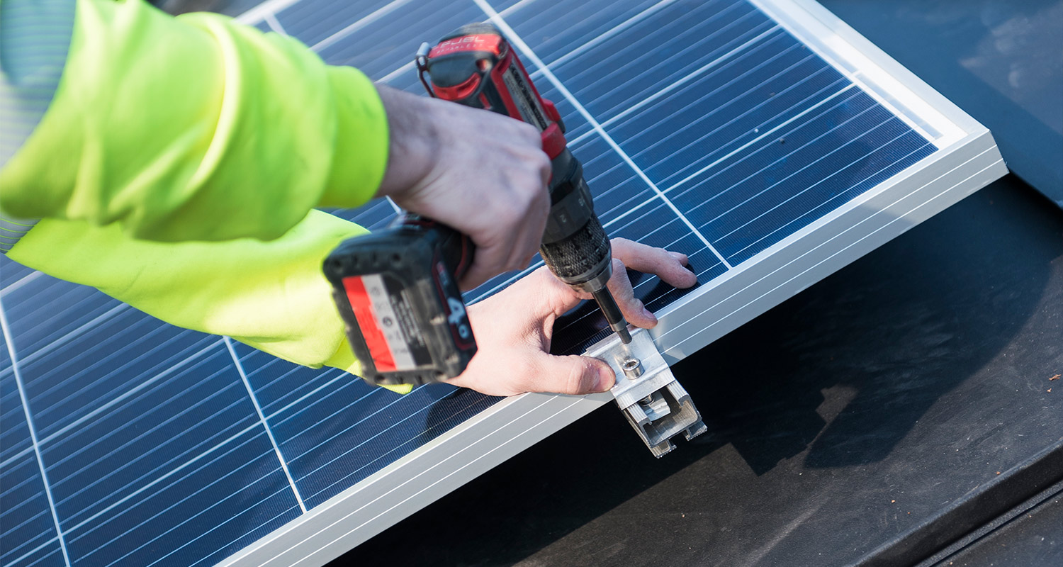 Asentaja neonkeltaisessa hupparissa poraa kiinnikettä siniseen aurinkopaneeliin.