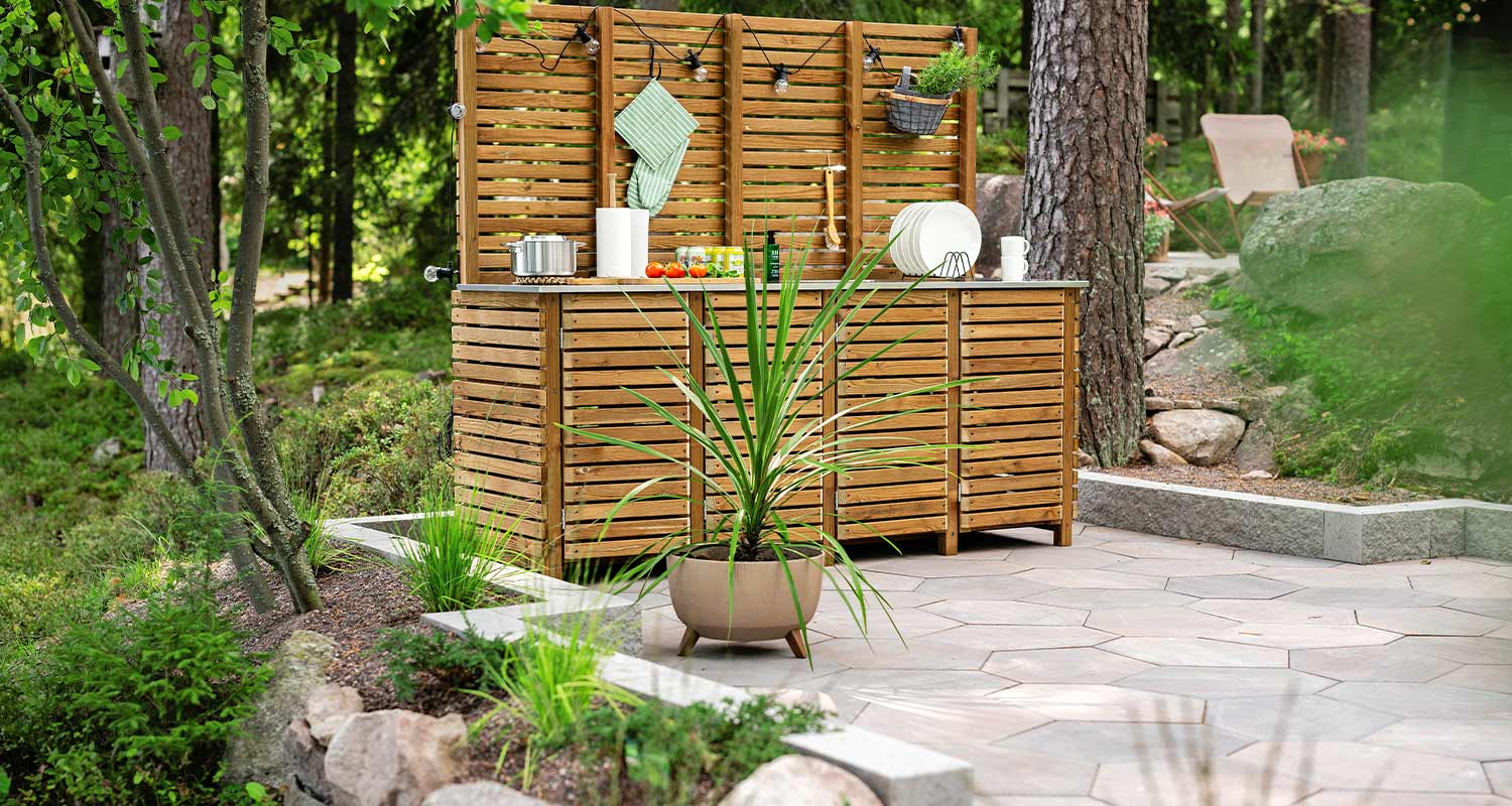 Puinen ulkokeittiö harmaalla laattaterassilla, ruukkuvehka ja vehreä metsänäkymä.