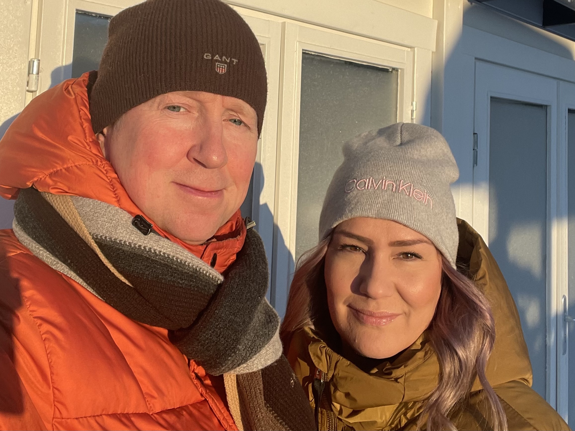 Oranssi- ja oliivinvihreät toppatakit ja pipot valkoista ovea vasten talvisessa auringossa.