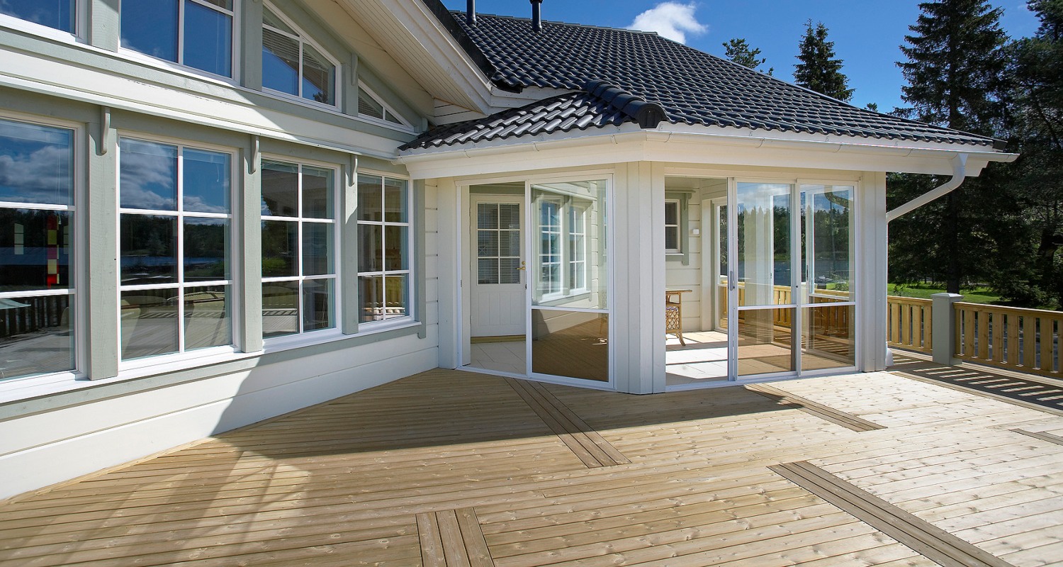 Vaalea hirsitalo mustalla tiilikatolla, lasikuisti ja suuri puuterassi järven rannassa