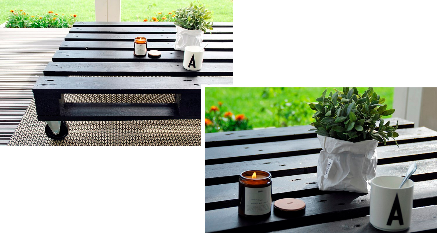 Musta puupallet-pöytä terassilla, palava kynttilä lasipurkissa, valkoinen A-muki ja ruukkukasvi.
