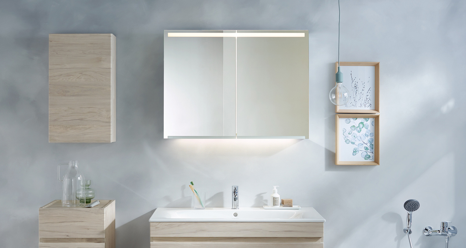 Harmaa kylpyhuone: vaaleat puukaapit, kasvikuvioiset taulut, valaistu peilikaappi ja pesuallas.