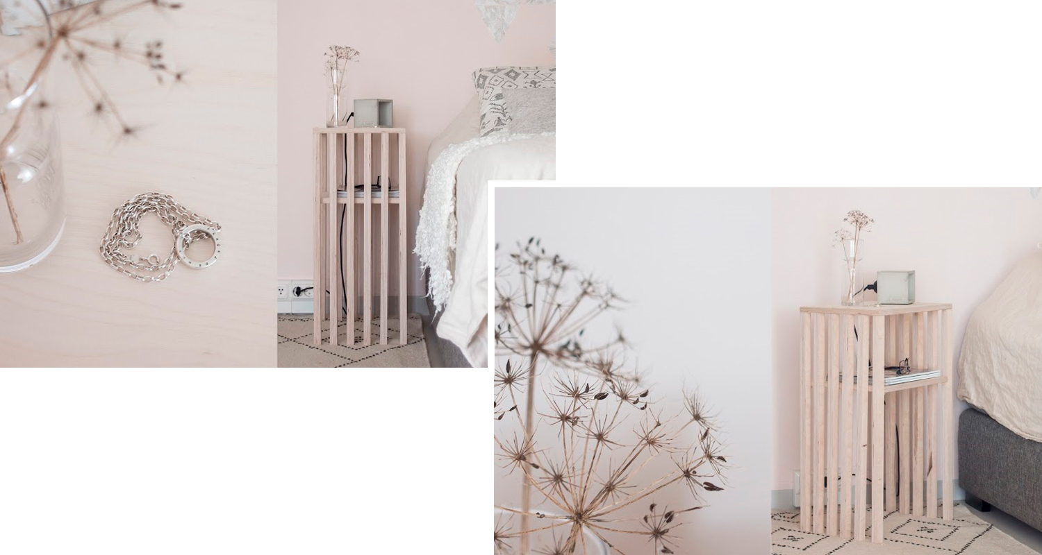 Vaalea puulankkupöytä sängyn vieressä vaaleanpunaisella seinällä, maljakossa kuiva kukka ja hopeaketju.