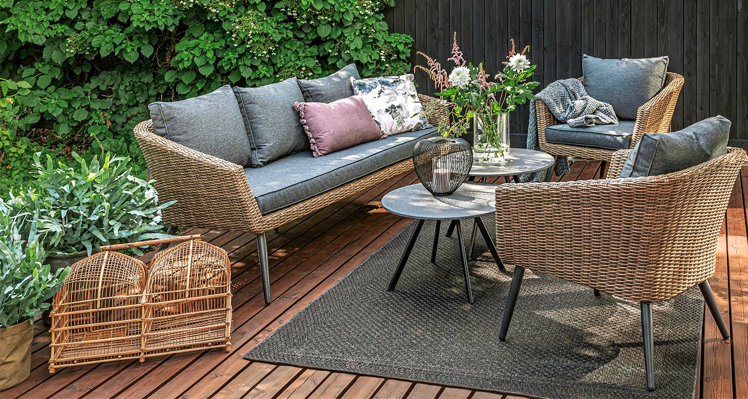 Rottinkinen terassiryhmä harmailla tyynyillä, pyöreillä pöydillä ja vihreillä kasveilla