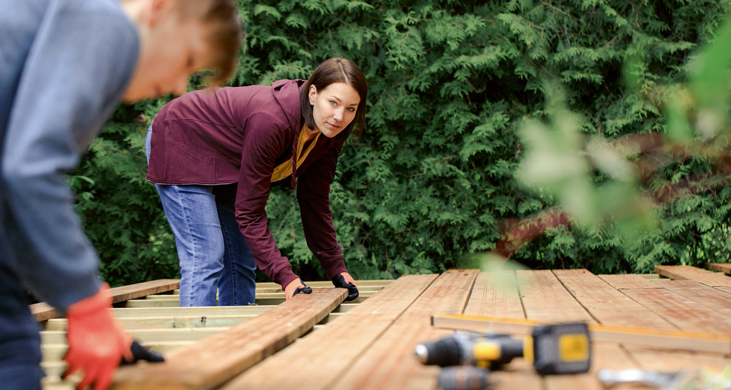 Kaksi ihmistä asentaa ruskeita terassilautoja vihreän pensasaidan edessä, porakone sivulla.