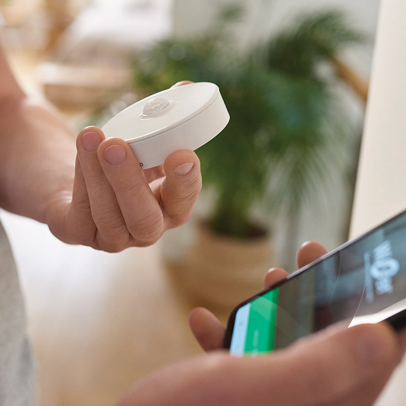 Käsi pitelee pientä valkoista pyöreää liiketunnistinta älypuhelimen edessä