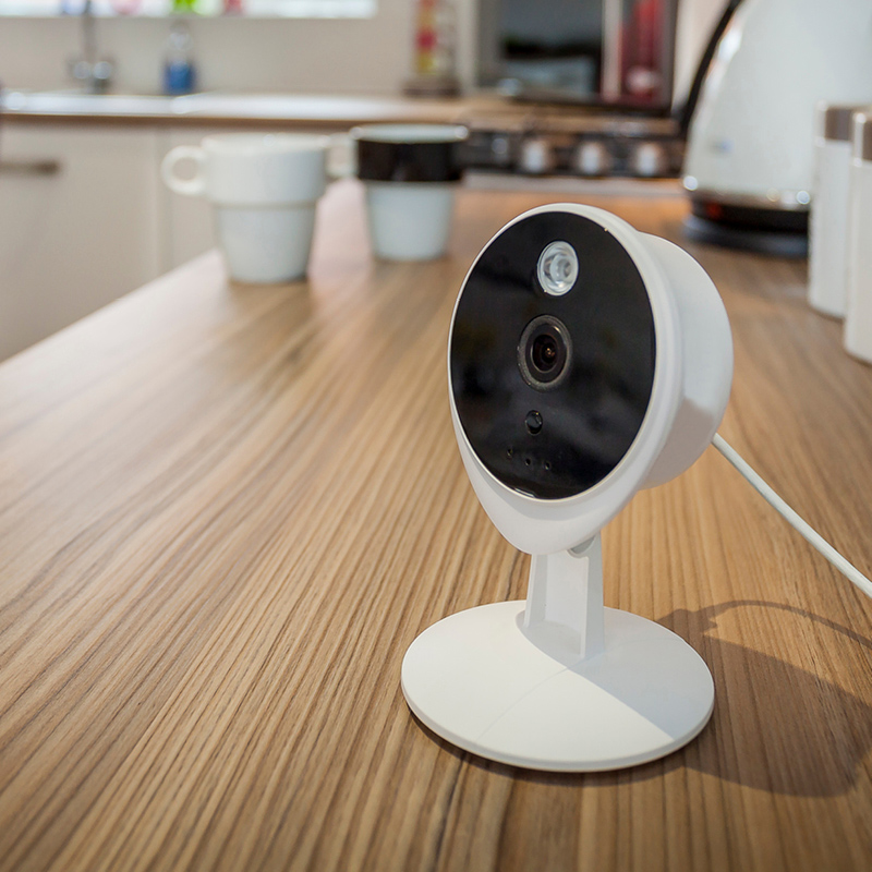 Valkoinen turvakamera mustalla etupaneelilla puupöydällä, keittiössä kahvikupit taustalla