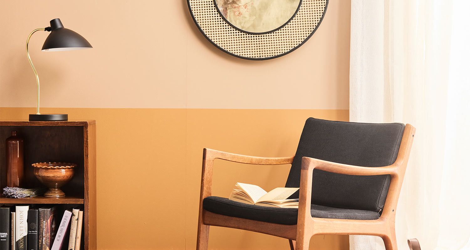 Puinen mustatuoli avoimella kirjalla oranssinkeltaista seinää vasten, hyllyllä lamppu ja peili.