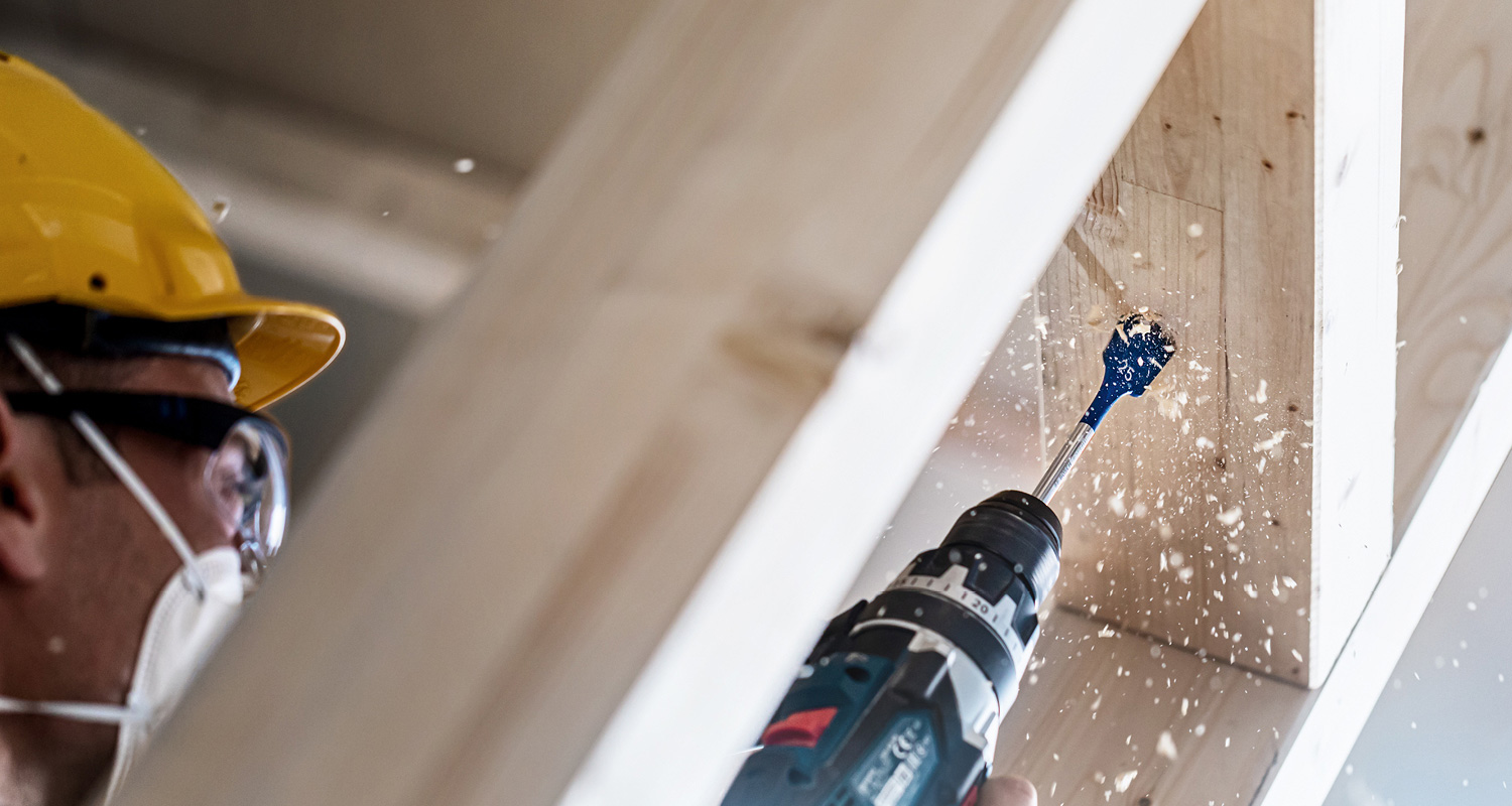 Rakennusmies keltakypärässä poraa alakaton puupalkkia sähköporalla, lastuja lentää.